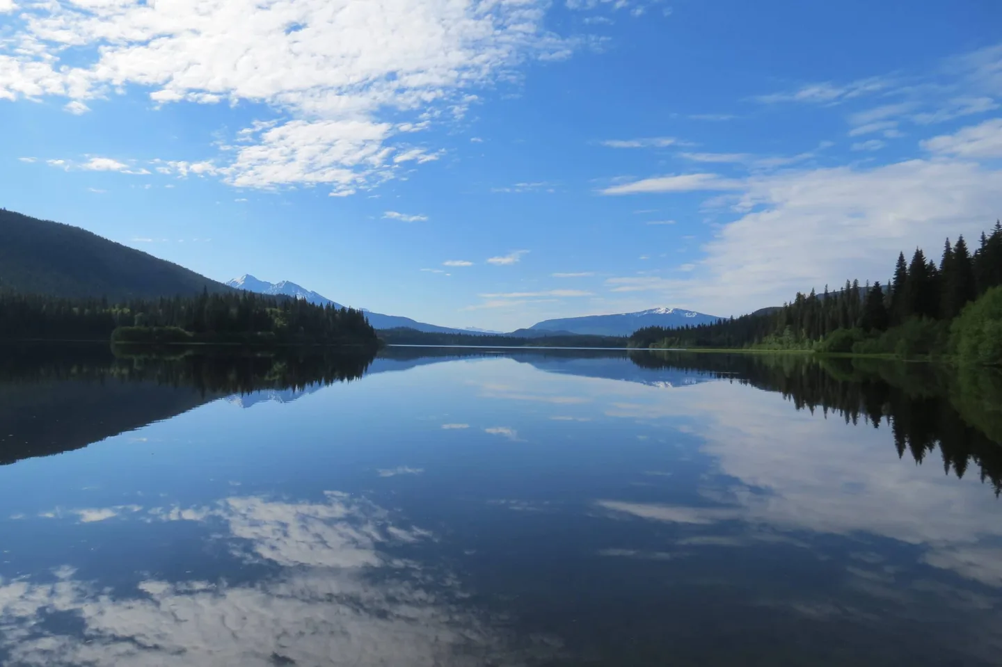 A wide channel of Bowron Lake running between forested hills under a bright cloudy sky